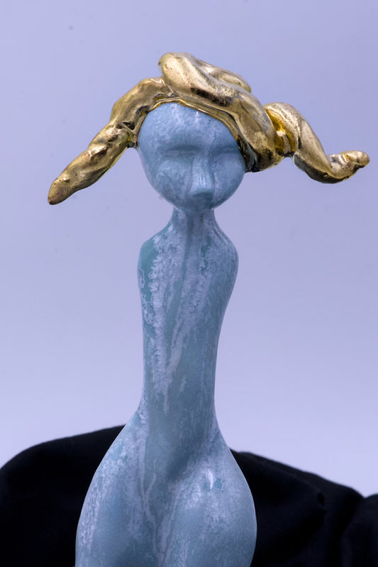 Blue ceramic figure with gold headpiece on a black surface against a light blue background